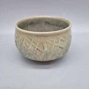 May include: A handmade ceramic bowl with a textured, crosshatch pattern. The bowl has a light beige exterior with subtle blue-green accents, and a smooth interior. The bowl is a round shape and is suitable for serving food.