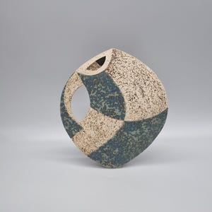 May include: A textured ceramic vase with a distinctive, geometric design. The vase has a square shape with a circular opening and a handle-like cutout. The colour scheme includes beige, speckled with brown, and sections of blue. The vase is a decorative art piece.