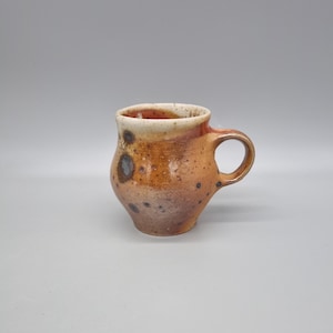 May include: A handcrafted ceramic mug with a rounded body and a handle. The mug has a speckled, earthy colour palette of brown, orange, and tan. The interior rim is a lighter tan colour. The mug is suitable for coffee, tea, or other beverages.