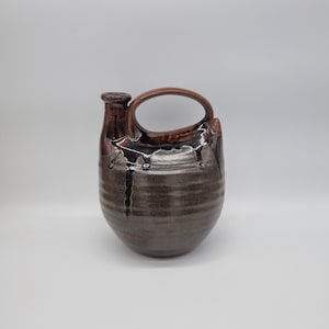 May include: A handcrafted ceramic jug with a dark brown glaze and a handle. The jug features a rounded body and a narrow neck with a spout. The glaze has a dripped effect, adding texture and visual interest to the piece.
