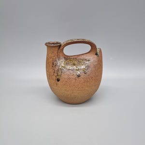 May include: A handmade, ceramic pitcher with a handle and spout. The pitcher has a rounded body and a textured, earthy brown and tan color scheme. The handle is arched, and the spout is small and cylindrical. The pitcher has a rustic, artisanal aesthetic.