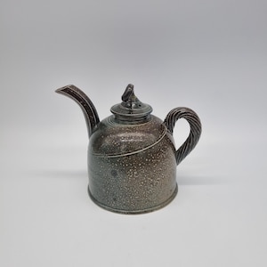 May include: A speckled brown ceramic teapot with a twisted handle and a curved spout. The lid has a small loop on top. The teapot has a dark brown rim and a light brown body. The teapot is a kitchenware item.