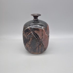 May include: A handmade ceramic vase with a dark brown and reddish-brown textured finish. The vase has a rounded body and a narrow neck. The design features a geometric pattern and a matte finish, suitable for home decor.