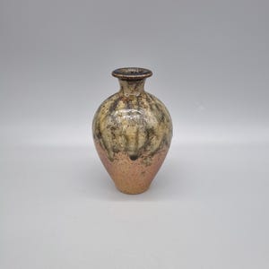 May include: A handcrafted ceramic vase with a rounded body and a narrow neck. The vase features a blend of colors, including beige, brown, and a copper-colored base. The surface has a textured appearance, adding to its unique aesthetic. This decorative vase is suitable for home decor.