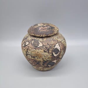 May include: A handmade ceramic jar with a lid. The jar is a mottled tan colour with dark brown and white abstract designs. The lid has a similar design. The jar has a rounded shape.