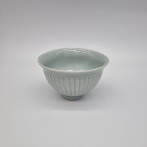 May include: A pale celadon ceramic bowl with vertical ribbed detailing. The bowl has a rounded shape with a slightly flared rim. The bowl is likely used for serving food or as a decorative piece.
