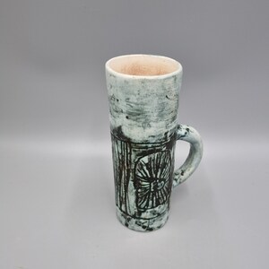 May include: A ceramic mug with a green and white glaze and a black design. The mug has a handle and is cylindrical in shape.