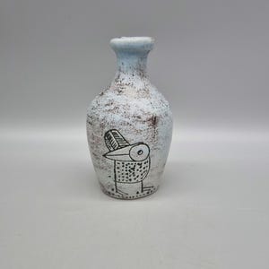 May include: A small, light blue ceramic vase with a stylised bird design in black. The vase has a distressed, aged look.