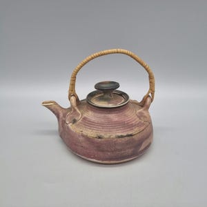 May include: A small, purple ceramic teapot with a woven wicker handle and a lid. The teapot has a rustic, handmade look.