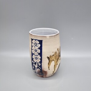 May include: A white ceramic mug with a blue and white floral design and a brown abstract design. The mug is slightly curved and has a rounded bottom.