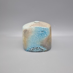 May include: A ceramic vase with a rounded top and a cylindrical body. The vase has a textured surface with blue, brown, and beige colours. A small, circular opening is visible on the top.