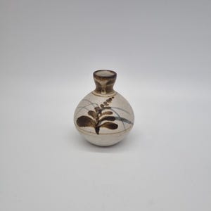 May include: A small, round ceramic vase with a narrow neck. The vase is off-white with brown leaf and line designs. The rim and neck are a darker brown. The vase is approximately 3 inches tall.