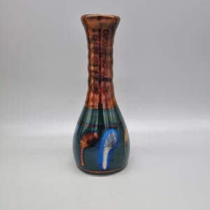 May include: A decorative vase with a dark green base and a copper-coloured neck. The vase has a bulbous shape with a long, textured neck. The base features blue and brown abstract designs. The vase is likely made of ceramic or glass.