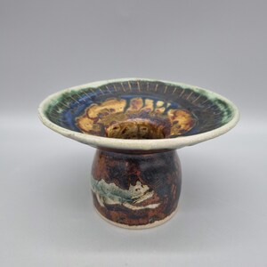 May include: A small ceramic pedestal with a wide, flat top and a round base. The pedestal is decorated with brown, green, and blue glaze, and has a white rim.