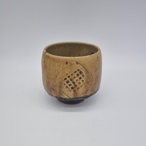 May include: A tan ceramic tea bowl with a textured surface and a diamond pattern carved into the side. The bowl has a rounded shape and a dark brown base. The interior is a lighter tan colour.