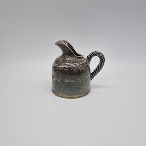 May include: A small, speckled ceramic pitcher with a dark brown and green glaze. The pitcher has a spout, a twisted handle, and a round base. The overall design is rustic and handmade, suitable for serving sauces or cream.