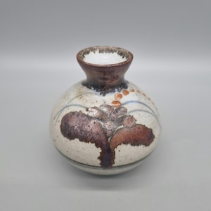 May include: A small, round, white ceramic vase with brown and purple floral designs. The vase has a slightly flared opening.