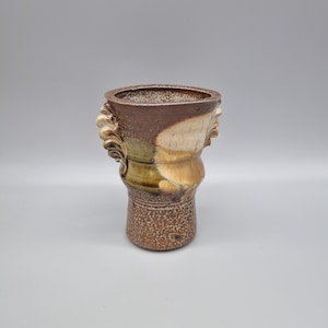 May include: A handcrafted ceramic vase with a textured surface and a unique design. The vase features a combination of brown, beige, and green hues. It has a wide opening and decorative handles on each side. The vase is approximately 8 inches tall.