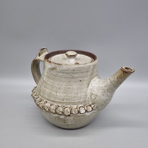 May include: A handcrafted ceramic teapot with a speckled, off-white glaze. The teapot features a brown rim, handle, and spout, and a decorative border around the base. The lid has a small knob. A unique piece for tea lovers.