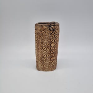 May include: A rectangular vase with a carved floral pattern. The vase is a warm brown colour and has a textured surface. The opening is rectangular. The vase has a rustic, handmade appearance, suggesting it is made from clay.