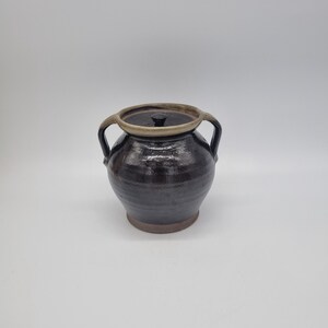 May include: A dark brown ceramic jar with a lid and two handles. The jar has a round body and a slightly flared base. The lid is a lighter brown colour. The jar is likely used for storage or decoration.