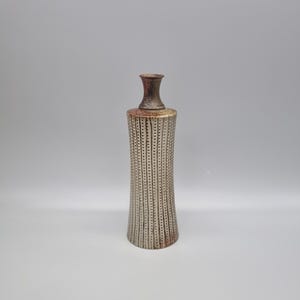 May include: A decorative vase with a narrow neck and a flared base. The vase has a textured, vertical striped pattern in a neutral tone, with a reddish-brown rim and neck. The vase is likely made of ceramic or pottery.