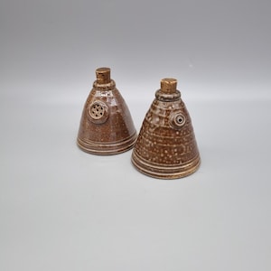 May include: Two brown ceramic bottle stoppers with cork tops. Each stopper has a conical shape with a textured surface and decorative details. One has a circular design with holes, the other has a small circular hole.