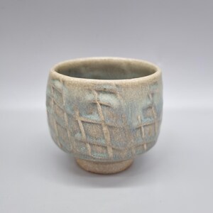 May include: A handcrafted ceramic cup with a textured, grid-like design. The cup has a rounded form and a small, circular base. The colour scheme includes shades of blue, beige, and brown, giving a rustic appearance. The cup is suitable for beverages.
