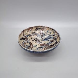 May include: Oval ceramic bowl with a cream interior and a blue rim. The bowl features a hand-painted design of fish and abstract shapes in blue and brown. A decorative piece for the home, ideal for serving.