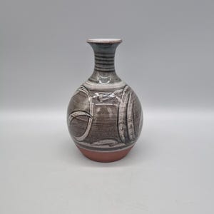 May include: A small, round, brown ceramic vase with white and grey painted designs. The vase has a narrow neck and a slightly flared lip.