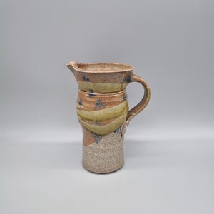 May include: A handmade ceramic pitcher with a spout and handle. The pitcher features a beige base, a tan middle section with green and blue accents, and a tan rim. The design includes a textured surface and a curved handle.