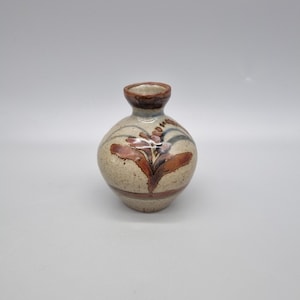 May include: A small, round ceramic vase with a narrow neck. The vase is beige with brown and reddish-brown floral designs. The rim and the base of the neck are brown. The vase has a bulbous body and a small opening.