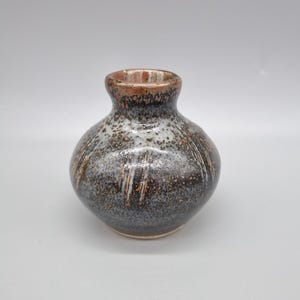 May include: A small, round ceramic vase with a narrow neck. The vase has a speckled, dark brown and grey glaze with streaks of white and orange. The rim is a darker brown colour.