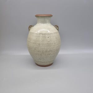 May include: A cream-coloured ceramic vase with a wide body, narrow neck, and two small handles. The vase has a textured surface with a brown rim and base. The design includes a horizontal band near the top and bottom.