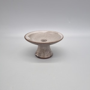 May include: A speckled ceramic pedestal bowl. It has a wide, shallow dish with a central hole, and a rounded base. The bowl is a neutral colour with a slightly textured surface. The design is simple and elegant, suitable for displaying small items.