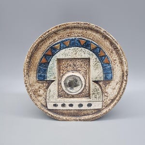 May include: A round, ceramic decorative plate with a textured, earthy brown rim. The center features a geometric design with blue, orange, and white accents. The plate has a circular indentation in the center.