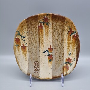 May include: A round, off-white ceramic plate with brown and green stripes and red flower designs. The plate has a small, brown, circular mark with a flower and the letter "M" in the centre.