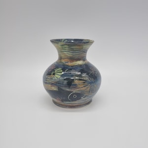 May include: A small, round ceramic vase with a narrow neck and flared rim. The vase has a dark blue background with painted details in green, brown, and white. The design includes a fish and other abstract shapes.