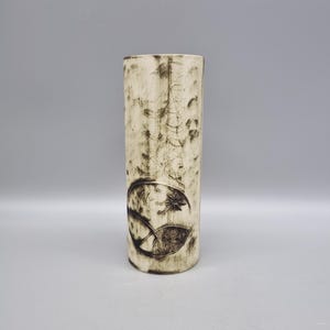 May include: A white ceramic vase with a cylindrical shape and a dark brown design of a stylised flower and leaves.