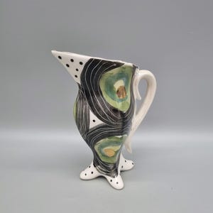 May include: A white ceramic jug with a black and green abstract design. The jug has a handle and three legs with black polka dots.