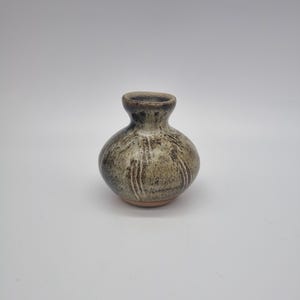 May include: A small, handcrafted ceramic vase with a rounded body and a narrow neck. The vase features a speckled, earthy color palette of brown, tan, and gray, with vertical streaks adding texture. The base is a lighter shade of brown.