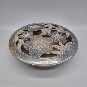 May include: A ceramic bowl with a floral design. The bowl is round with a blue and brown colour scheme. The top of the bowl features cut-out flowers and leaves. The bowl is likely used for decorative purposes or serving food.
