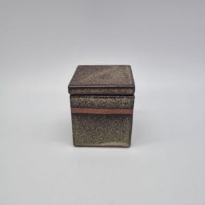 May include: A square ceramic box with a lid, featuring a speckled dark brown and tan glaze. The exterior surface displays a distinct horizontal band of unglazed reddish-brown clay circling the upper portion of the container body.