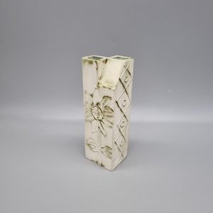 May include: An off-white ceramic vase with a rectangular shape, featuring a floral design and diamond pattern. The vase has a square opening and is decorated with a raised sunflower and leaf motif. The edges have a green tint.