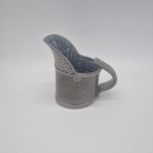 May include: A grey ceramic jug with a textured surface and a handle. The jug has a spout and a decorative rim. The interior is a darker grey. The jug is likely used for serving liquids.