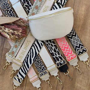 Könnte beinhalten: Eine Sammlung von bunten und gemusterten verstellbaren Riemen für Taschen. Die Riemen sind aus gewebtem Stoff und haben goldfarbene Metallbeschläge. Die Riemen sind fächerförmig auf einer Holzoberfläche angeordnet. Die Riemen haben verschiedene Muster, darunter Zebra- und Leopardenmuster sowie geometrische Designs.