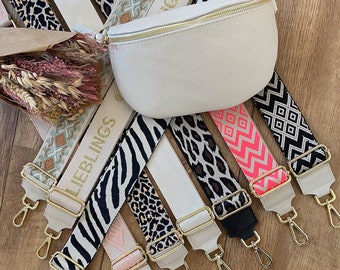 Puede incluir: Una colección de correas ajustables coloridas y estampadas para bolsos. Las correas están hechas de tejido tejido y tienen herrajes de metal dorado. Las correas están dispuestas en forma de abanico sobre una superficie de madera. Las correas tienen diferentes patrones, incluyendo estampado de cebra, estampado de leopardo y diseños geométricos.
