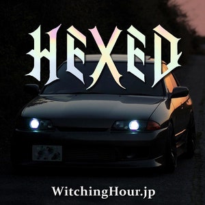 Puede incluir: Un coche negro con la palabra "HEXED" en letras iridiscentes en el parabrisas. El coche tiene las luces encendidas y el texto "WitchingHour.jp" está en la parte inferior de la imagen.