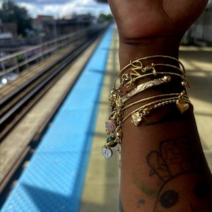 May include: A collection of gold bangles and charm bracelets adorns a wrist. The bracelets feature various charms, including flowers, hearts, and initials. The background shows a blue platform and train tracks.