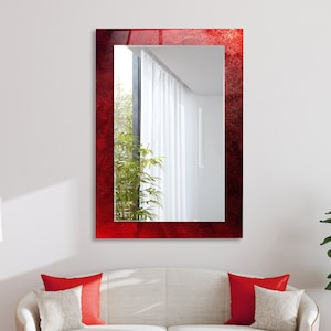K&ouml;nnte beinhalten: Ein roter Spiegel mit einem strukturierten Rahmen h&auml;ngt an einer wei&szlig;en Wand &uuml;ber einem wei&szlig;en Sofa mit roten Kissen. Der Spiegel spiegelt einen Raum mit einem Fenster und wei&szlig;en Vorh&auml;ngen wider.